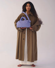Load image into Gallery viewer, a model wearing a trenchcoat standing against a neutral background holding a bundle of lavender andthe Ölend Mini Ona Soft Bag in lilac
