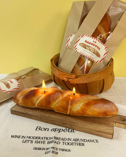 The Wednesday Co French Baguette Candle on a bread board styled in front of more candles in bread bags in a basket in the background 