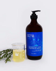 ALTR Hand & Body Wash in Eastern Red Cedar sitting next to a beaker of liquid soap in front of a cedar branch on a neutral background