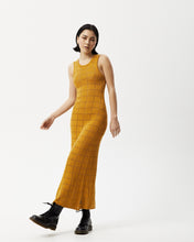Load image into Gallery viewer, the Afends Women&#39;s Femme Dress in Mustard on a model standing posing with her left leg across her body and her right arm out to the side 
