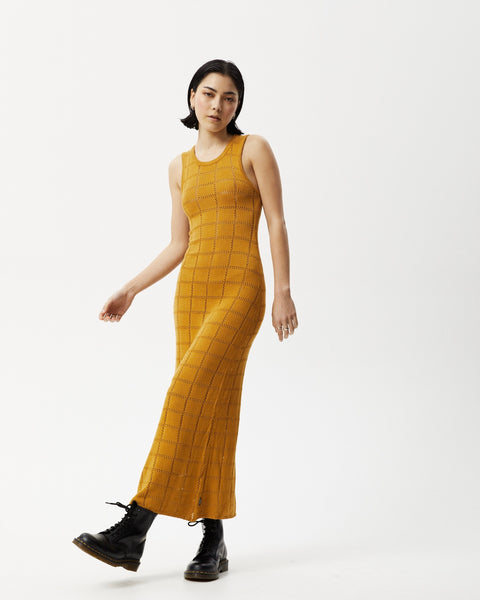 the Afends Women's Femme Dress in Mustard on a model standing posing with her left leg across her body and her right arm out to the side 