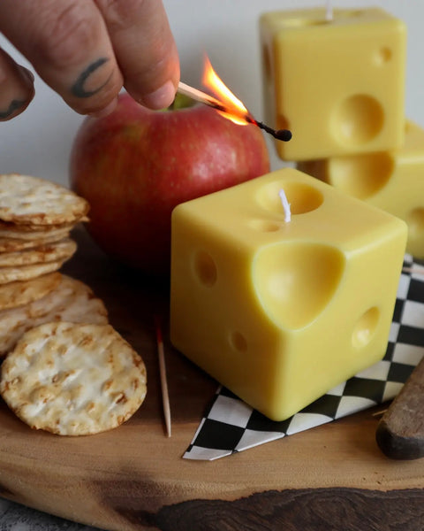 close up of a hand lighting the Drop Dead Swiss Cheese Cube Candle with a match on a charcuterie board
