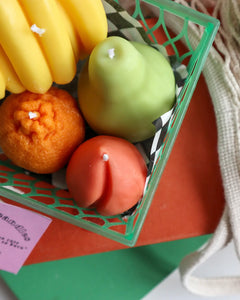 the Drop Dead Fruit Basket Candles sitting on a stack of books beside a net bag