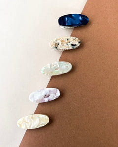 five oval Hair Clips in different colours line up on a diagonal line on top of a half brown half cream coloured surface