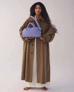 a model wearing a trenchcoat standing against a neutral background holding a bundle of lavender andthe Ölend Mini Ona Soft Bag in lilac