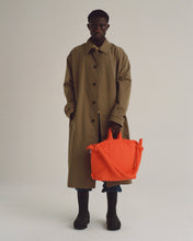 Load image into Gallery viewer, a model wearing a trenchcoat posing with the Ölend Ona Soft Bag in coral in his left hand and his right hand in his pocket
