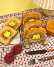 Load image into Gallery viewer, The Wednesday Co Buttered Toast Candles in a toast stand on a plaid tablecloth beside a plate of toast candles
