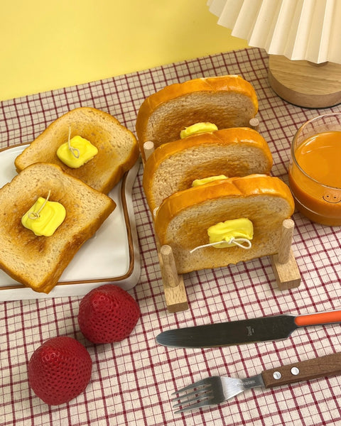 The Wednesday Co Buttered Toast Candles in a toast stand on a plaid tablecloth beside a plate of toast candles