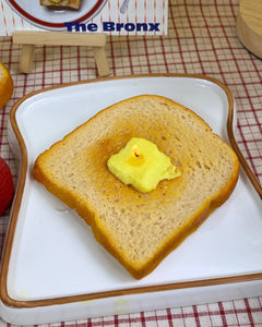 The Wednesday Co Buttered Toast Candle sitting on a white plate on a plaid tablecloth