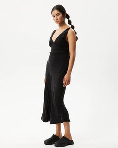 the Afends Women's Gemma Dress in Black on a model posing on a side angle in front of a white background staring into the camera