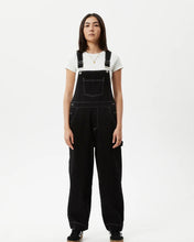Load image into Gallery viewer, the Afends Women&#39;s Louis Overalls in Washed Black on a model standing against a white background staring straight in to the camera
