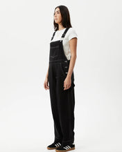 Load image into Gallery viewer, side angled view of the the Afends Women&#39;s Louis Overalls in Washed Black on a model standing against a white background
