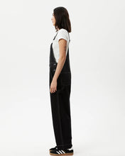Load image into Gallery viewer, side view of the the Afends Women&#39;s Louis Overalls in Washed Black on a model standing against a white background
