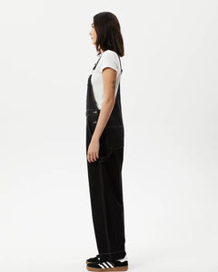 side view of the the Afends Women's Louis Overalls in Washed Black on a model standing against a white background
