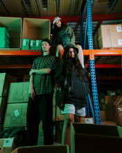 Load image into Gallery viewer, three models posing in a warehouse, two standing on the ground in front and one in the background sitting up high on a shelf
