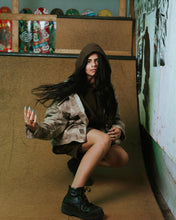 Load image into Gallery viewer, the Taikan Shirt Jacket in Abstract Camo worn by a model posing in a kneeling position flipping her long dark hair with her right hand
