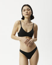 Load image into Gallery viewer, the Afends Women&#39;s Lolly Brief in Black on a model standing posing with her hands in front of her stomach
