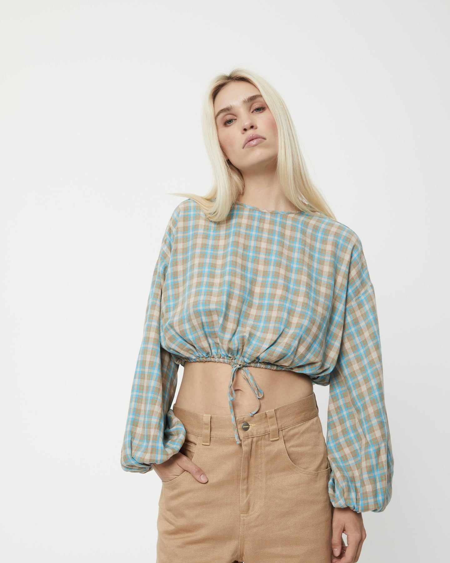 the Afends Women's Millie Blouse in Tan Check on a model posing with her hand in her pocket