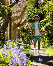 Load image into Gallery viewer, Poplin &amp; Co Men&#39;s Camp Shirt in Banana Bunch worn by a model walking in a lush green park filled with trees and purple flowers on a sunny day
