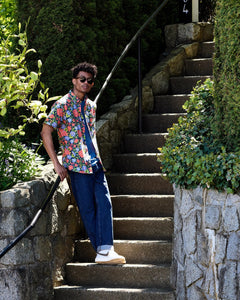 the Poplin & Co Men's Short Sleeve Printed Shirt in Hibiscus worn by a model posing against a railing in a stone stairway also wearing jeans, white nike sneakers and sunglasses