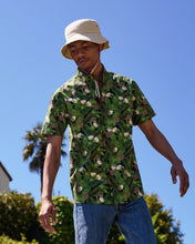 Load image into Gallery viewer, the Poplin &amp; Co Men&#39;s Short Sleeve Printed Shirt in Coconut worn by a model looking down toward his right and standing with his arms out outside against a blue sky and a palm tree 

