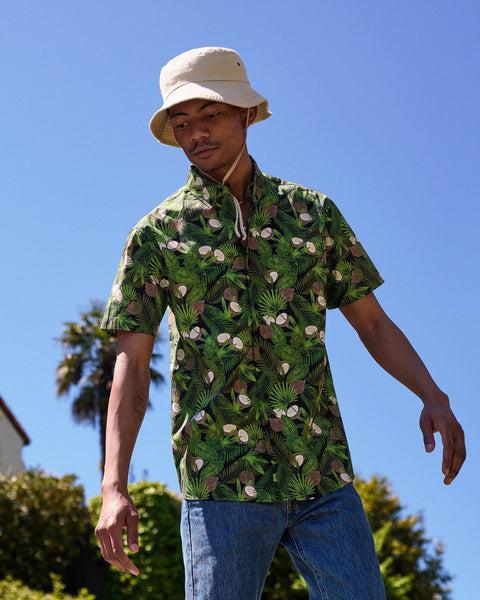 the Poplin & Co Men's Short Sleeve Printed Shirt in Coconut worn by a model looking down toward his right and standing with his arms out outside against a blue sky and a palm tree 