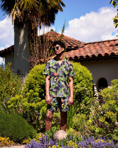 the Poplin & Co Men's Camp Shirt in Tropical Birds worn by a model standing in a lush garden with a spanish style house and palm tree in the background