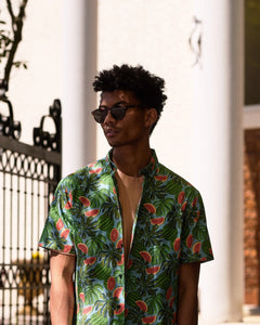 the Poplin & Co Men's Short Sleeve Printed Shirt in Watermelon worn open over a t-shirt by a model standing looking to the side with an iron gate in the background