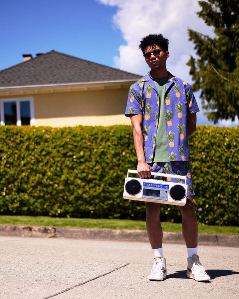 the Poplin & Co Men's Camp Shirt in Wild Pineapple worn by a model standing on a sidewalk in front of a house and a green hedge holding a boom box in one hand