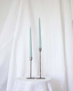 a pair of Socco Designs Taper Candles in light blue in two tapers of varying heights against a white sheet background